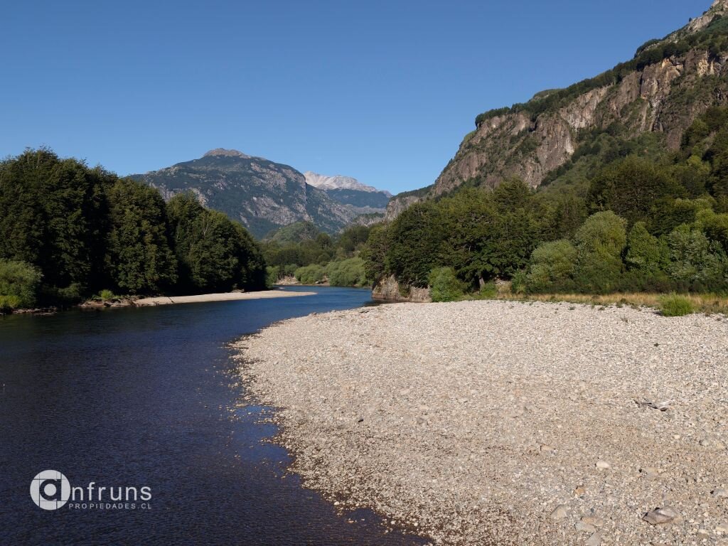Wide river with a rocky pebble beach and forested banks under a clear blue sky in Coyhaique.